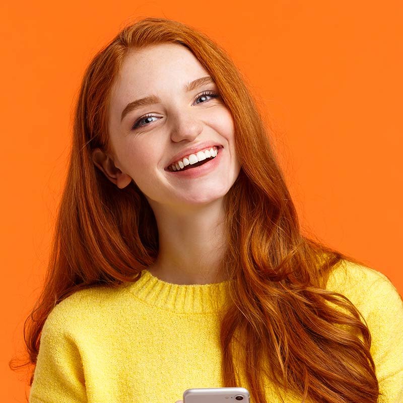 Girl searching gifts for holidays online. Carefree happy redhead female student using smartphone app, laughing joyfully, holding mobile phone, chatting or browsing social networks, orange background.
